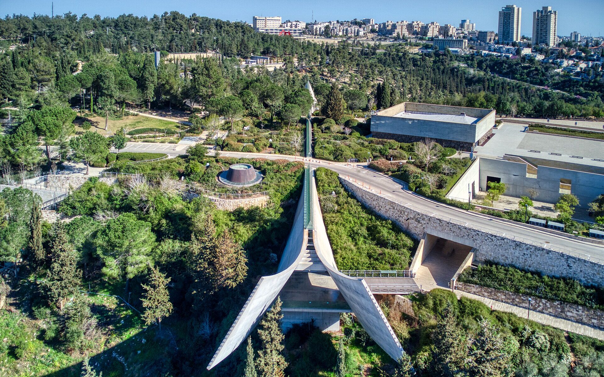 Holocaust History Museum at Yad Vashem seen from above, its concrete prism splaying open over a wooded valley, with the skylight ridge running back through the hillside