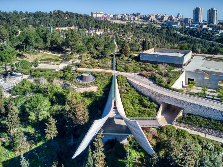 Holocaust History Museum at Yad Vashem seen from above, its concrete prism splaying open over a wooded valley, with the skylight ridge running back through the hillside