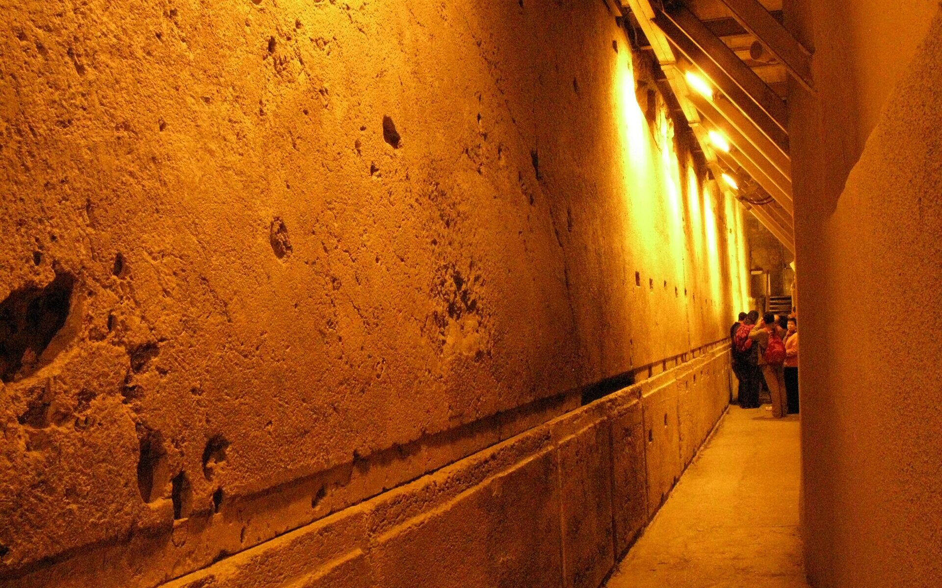 Huge Herodian limestone blocks of the Western Wall lit by warm lamps along a narrow underground passage, with a small group of visitors at the far end