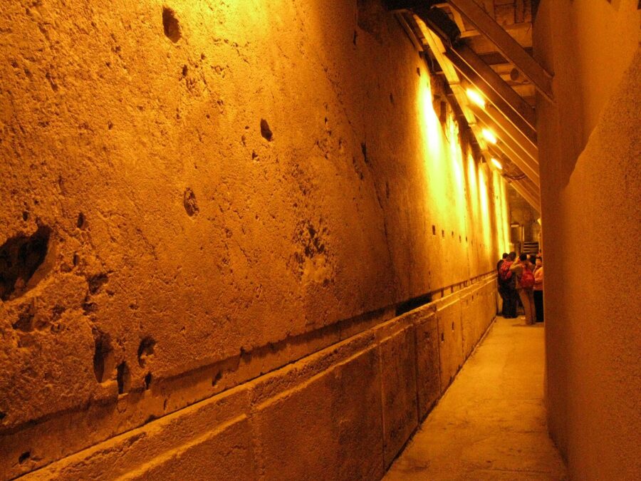 Huge Herodian limestone blocks of the Western Wall lit by warm lamps along a narrow underground passage, with a small group of visitors at the far end