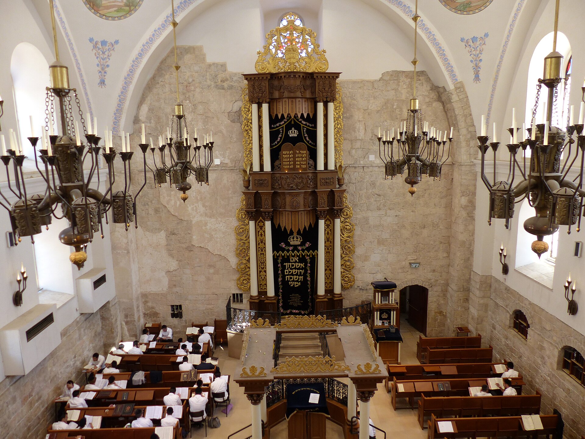 The Hurva Synagogue's prayer hall seen from the gallery, with a towering gilded Torah ark, brass chandeliers and men studying at wooden pews below