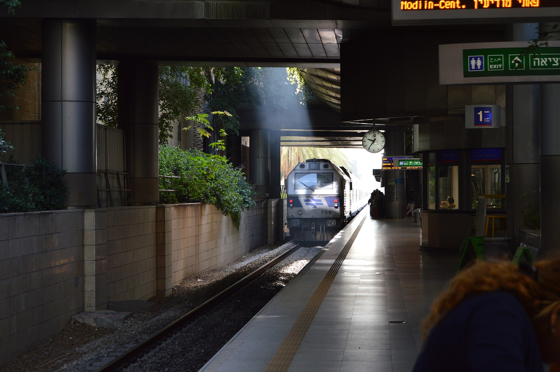 An Israel Railways locomotive pulls into platform 1 at Ben Gurion Airport station, with a platform clock, exit signs and a departure board overhead
