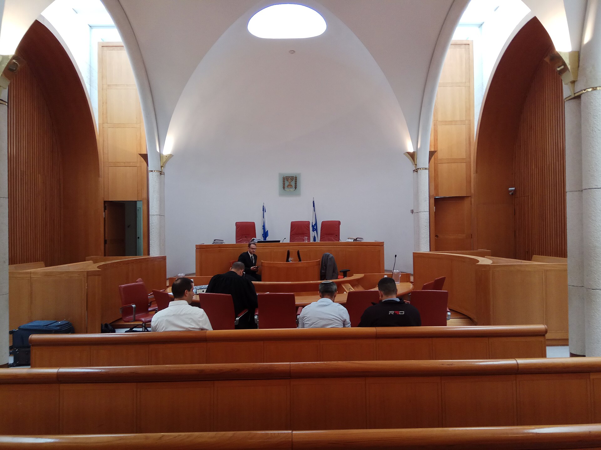 A hearing hall in Israel's Supreme Court with wood-panelled walls, red judges' chairs beneath the state emblem, flags, and lawyers seated at the front