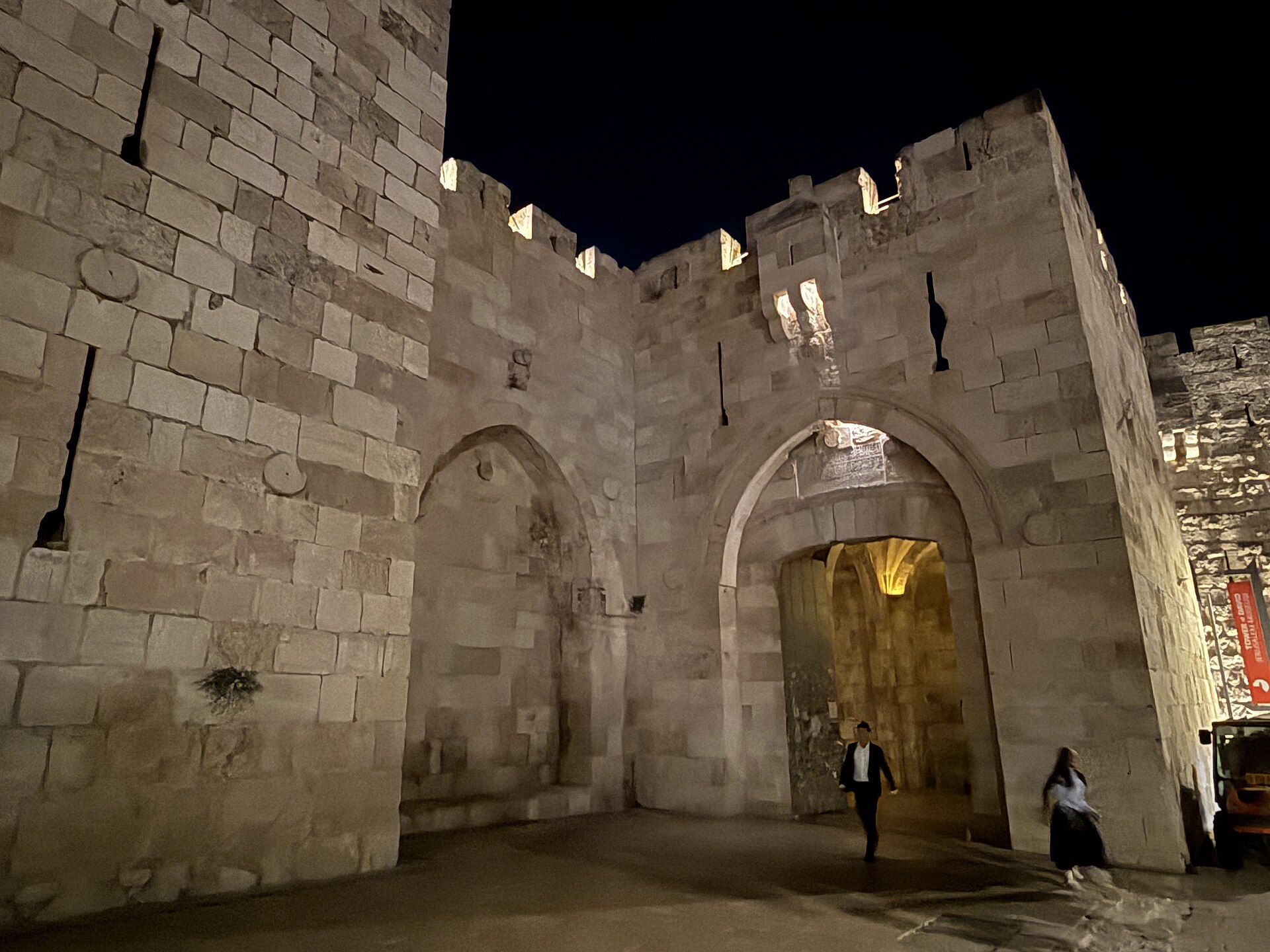 Jaffa Gate floodlit at night, its pointed-arch doorway glowing warmly beneath the inscription panel, beside a blind arch and crenellated walls, with two pedestrians passing