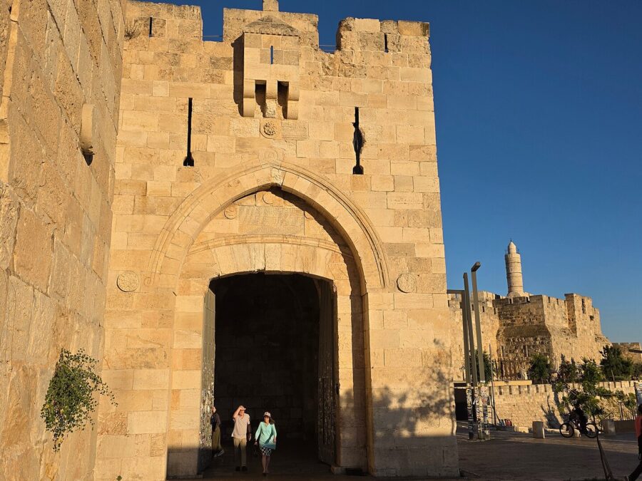 Jaffa Gate's pointed arch and Ottoman inscription panel glowing in evening sun, with visitors stepping out of the gateway and the Tower of David minaret beyond