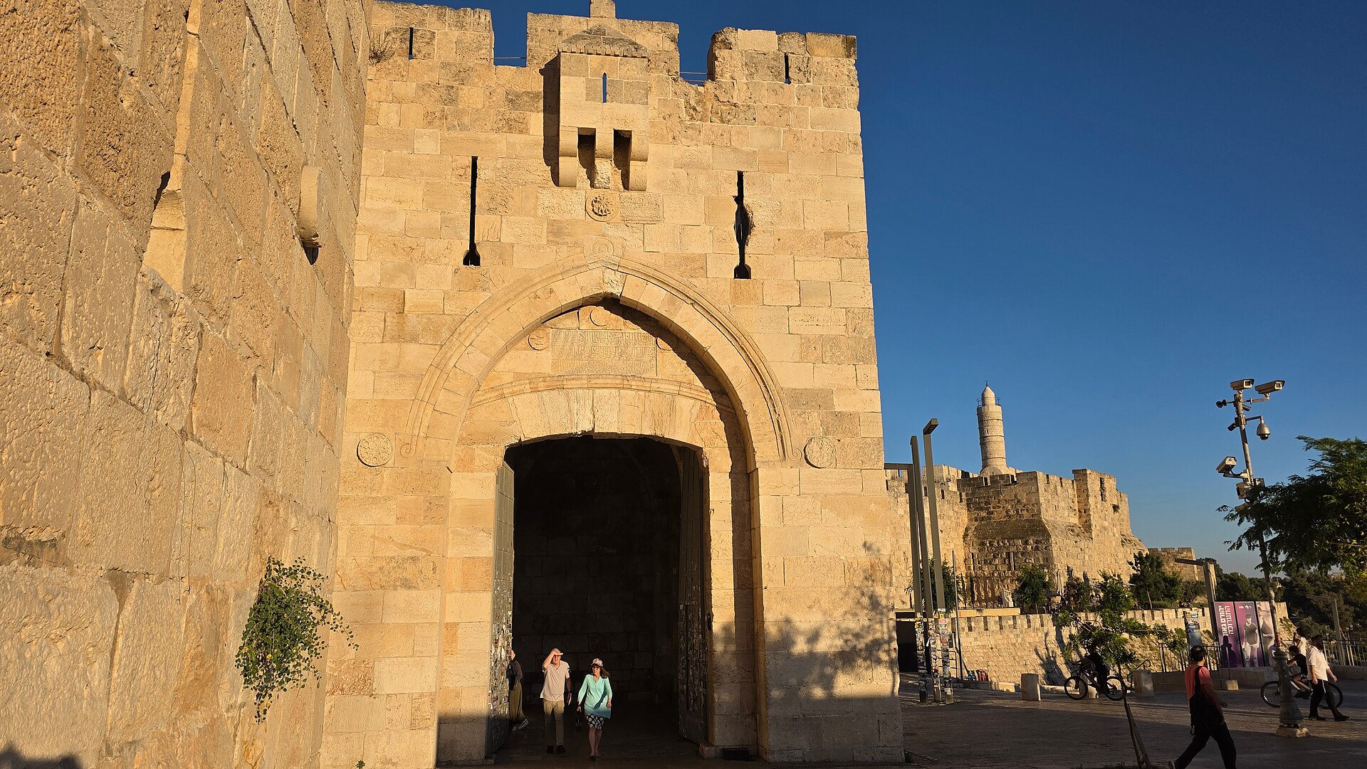 Jaffa Gate's pointed arch and Ottoman inscription panel glowing in evening sun, with visitors stepping out of the gateway and the Tower of David minaret beyond