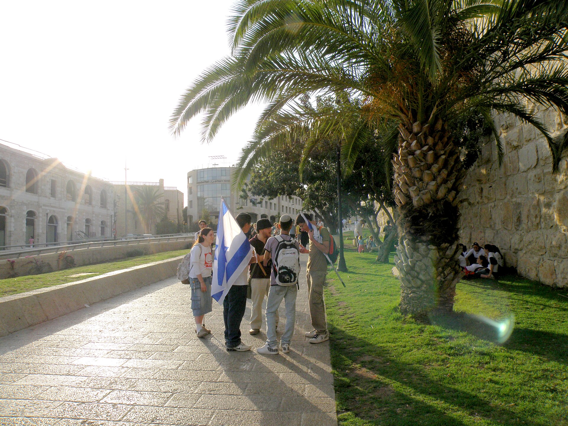 Young people carrying Israeli flags walking along a paved path beneath palm trees beside the Old City walls on Jerusalem Day