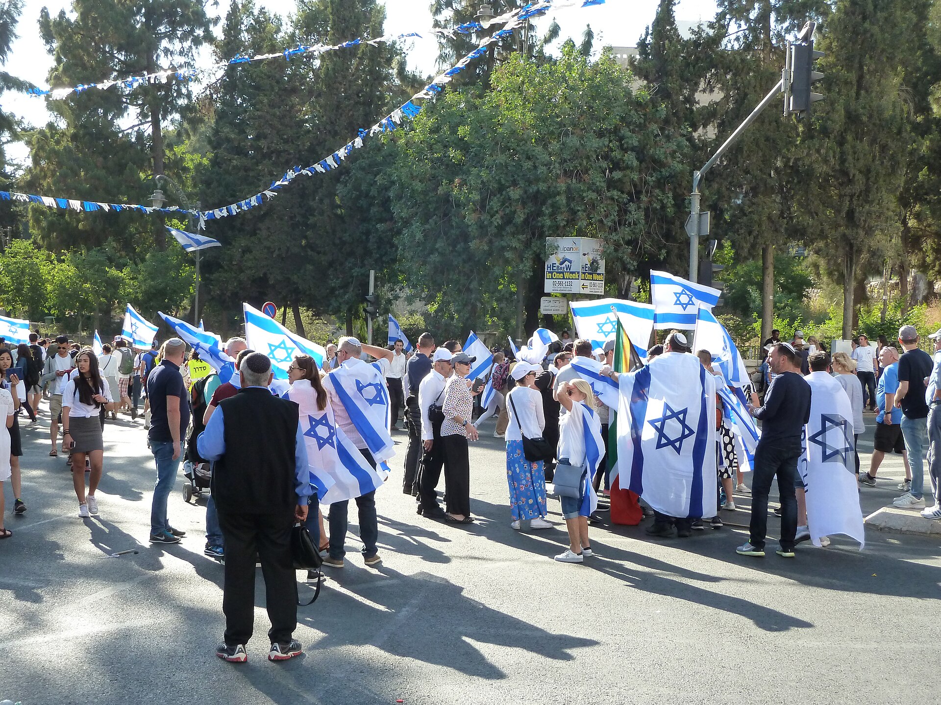 Crowds of marchers wrapped in and waving Israeli flags gathering on a tree-lined West Jerusalem street under strings of blue and white bunting