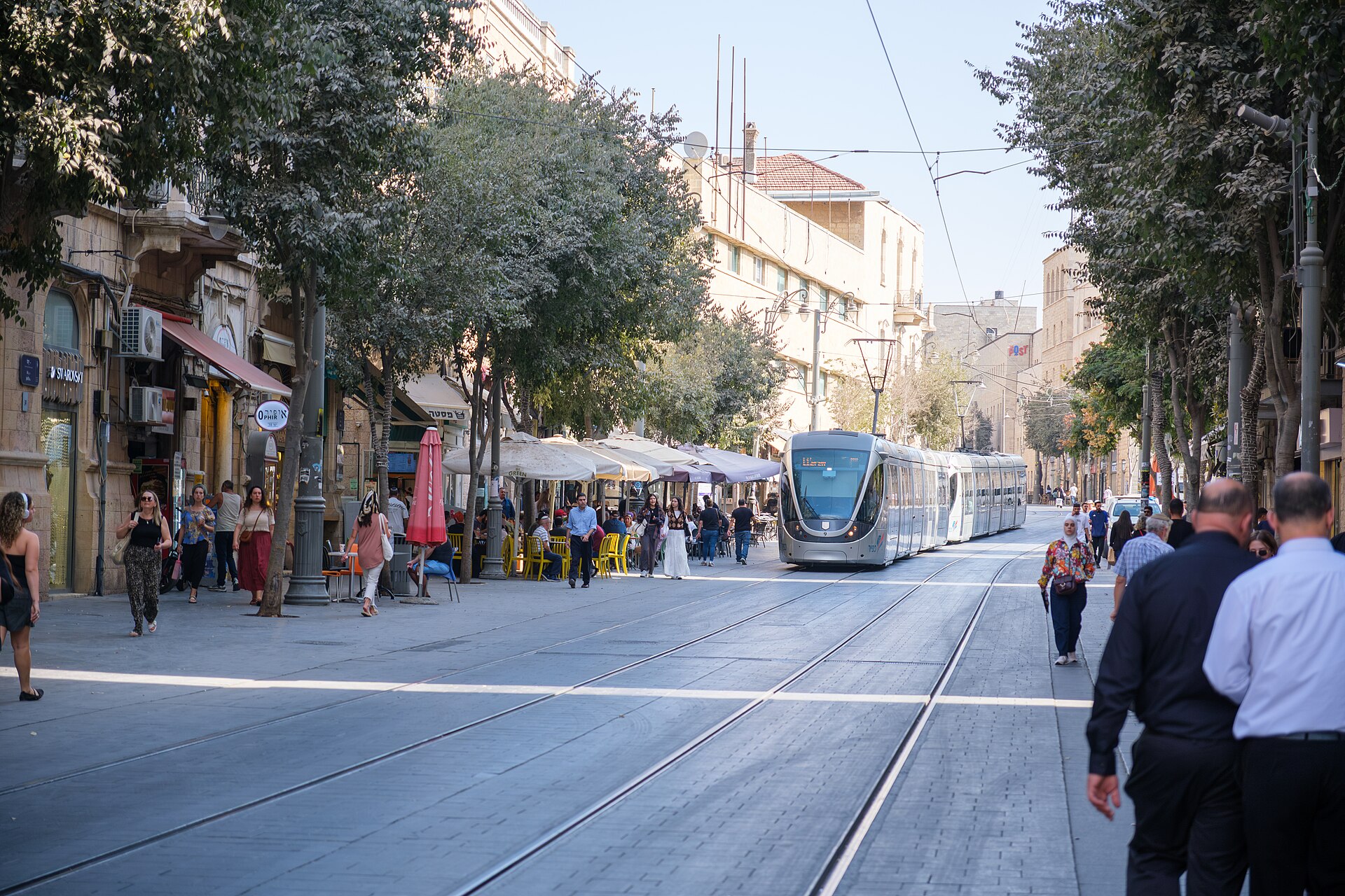 A silver Jerusalem light rail tram gliding along tree-lined, car-free Jaffa Road past café tables and pedestrians