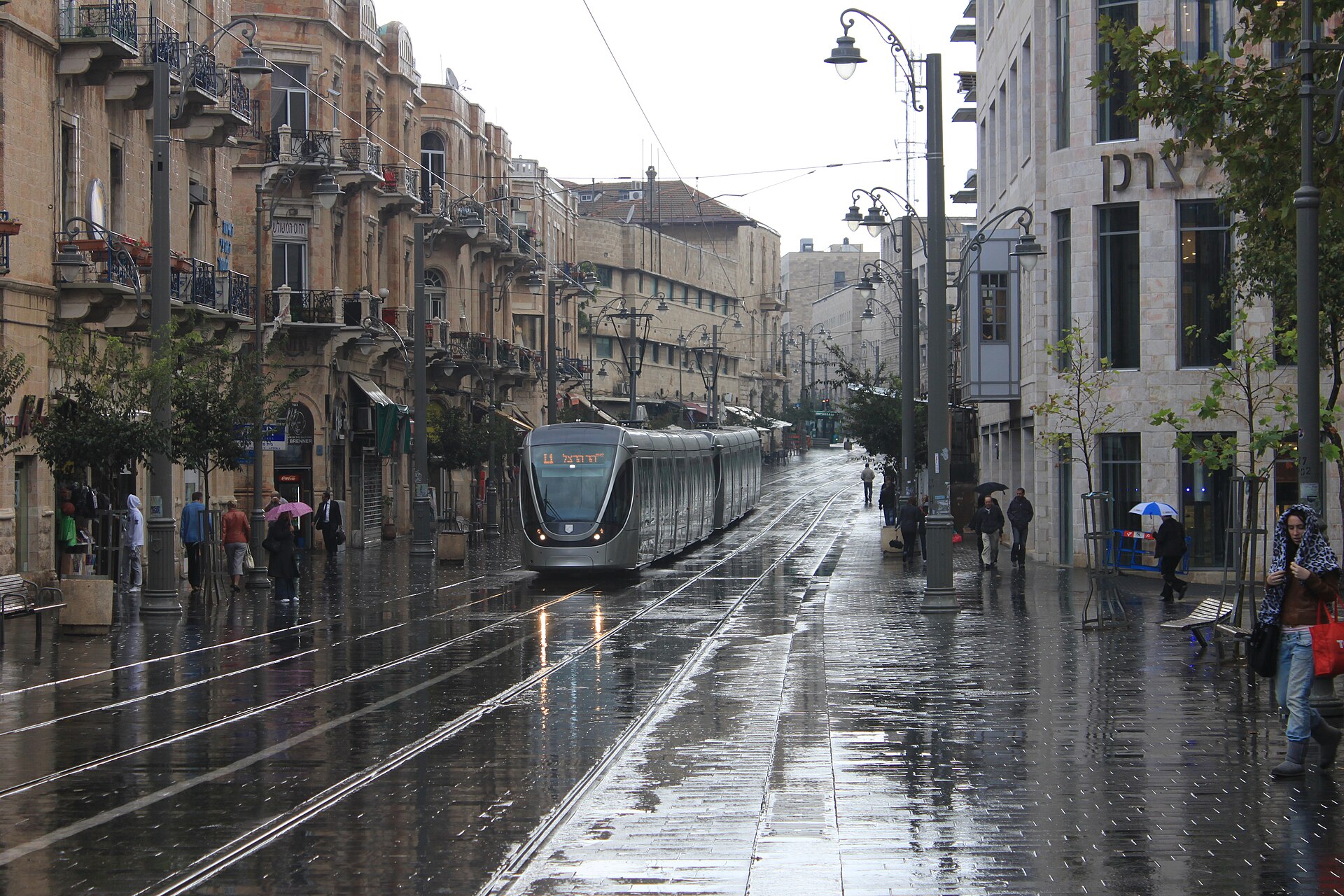 A Jerusalem Light Rail tram travels along rain-soaked Jaffa Road at Zion Square, its tracks and paving shining, as pedestrians hurry past under umbrellas
