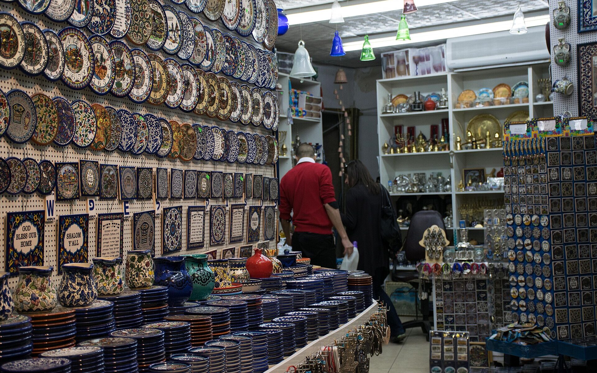 Jerusalem Old City souvenir shop with walls of hand-painted ceramic plates and tiles, stacked blue-and-white dishes, brass menorahs, glass bells and racks of magnets