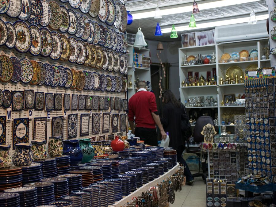 Jerusalem Old City souvenir shop with walls of hand-painted ceramic plates and tiles, stacked blue-and-white dishes, brass menorahs, glass bells and racks of magnets
