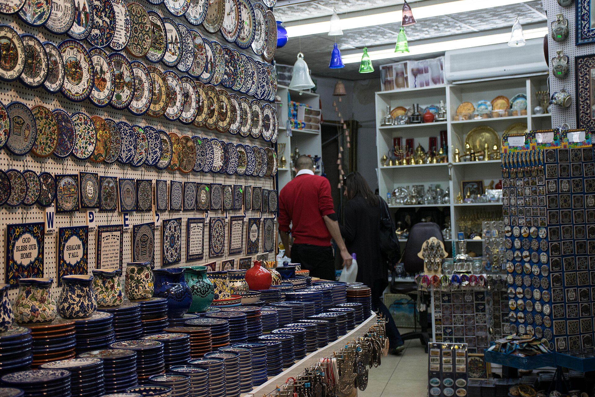 Jerusalem Old City souvenir shop with walls of hand-painted ceramic plates and tiles, stacked blue-and-white dishes, brass menorahs, glass bells and racks of magnets