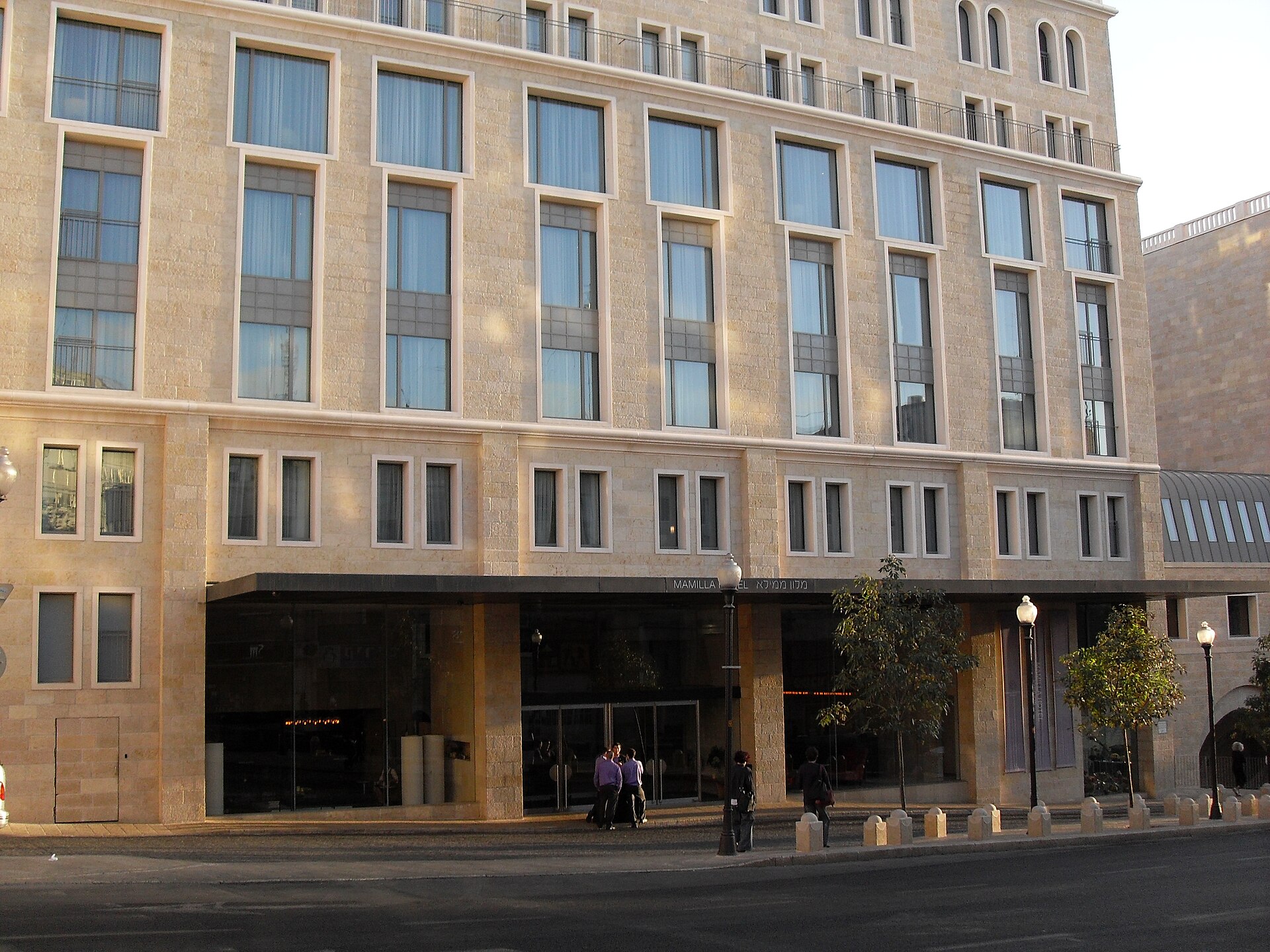 Jerusalem-stone façade of the Mamilla Hotel with rows of tall windows and a glass-fronted lobby beneath a canopy signed Mamilla Hotel in English and Hebrew