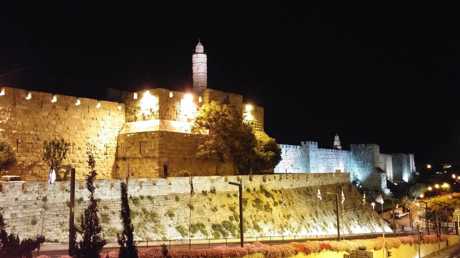 Jerusalem's Tower of David and western Old City wall floodlit at night, the citadel's minaret glowing gold as the crenellated wall runs on towards Mount Zion
