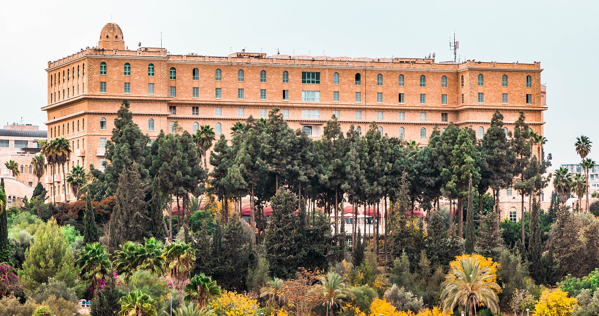 The King David Hotel's long limestone façade with a small corner dome and red terrace awnings, rising above cypresses, palms and flowering trees in Jerusalem