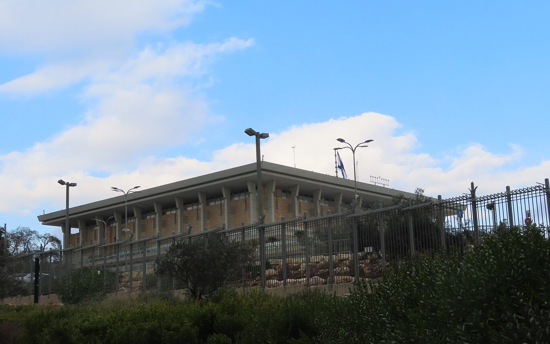 The Knesset building in Jerusalem, a flat-roofed stone block ringed by tall square columns, with an Israeli flag on the roof, seen from behind its security fence