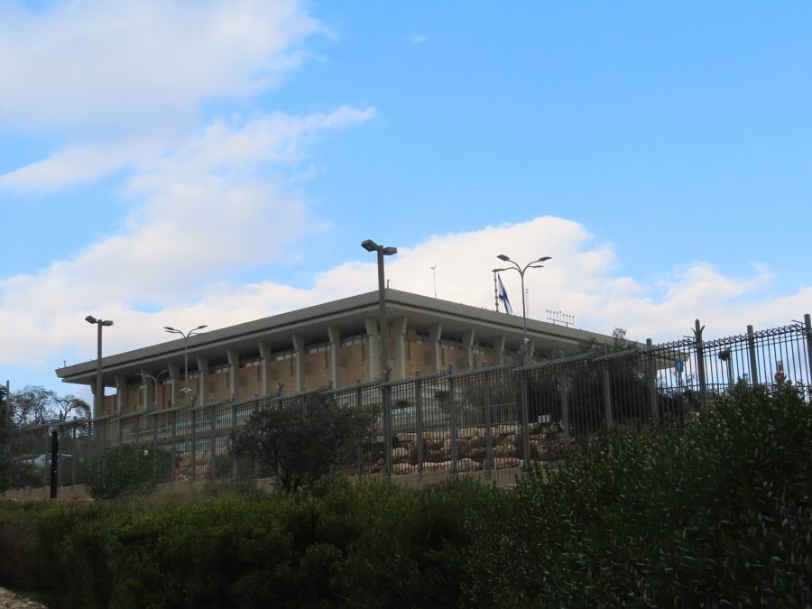 The Knesset building in Jerusalem, a flat-roofed stone block ringed by tall square columns, with an Israeli flag on the roof, seen from behind its security fence