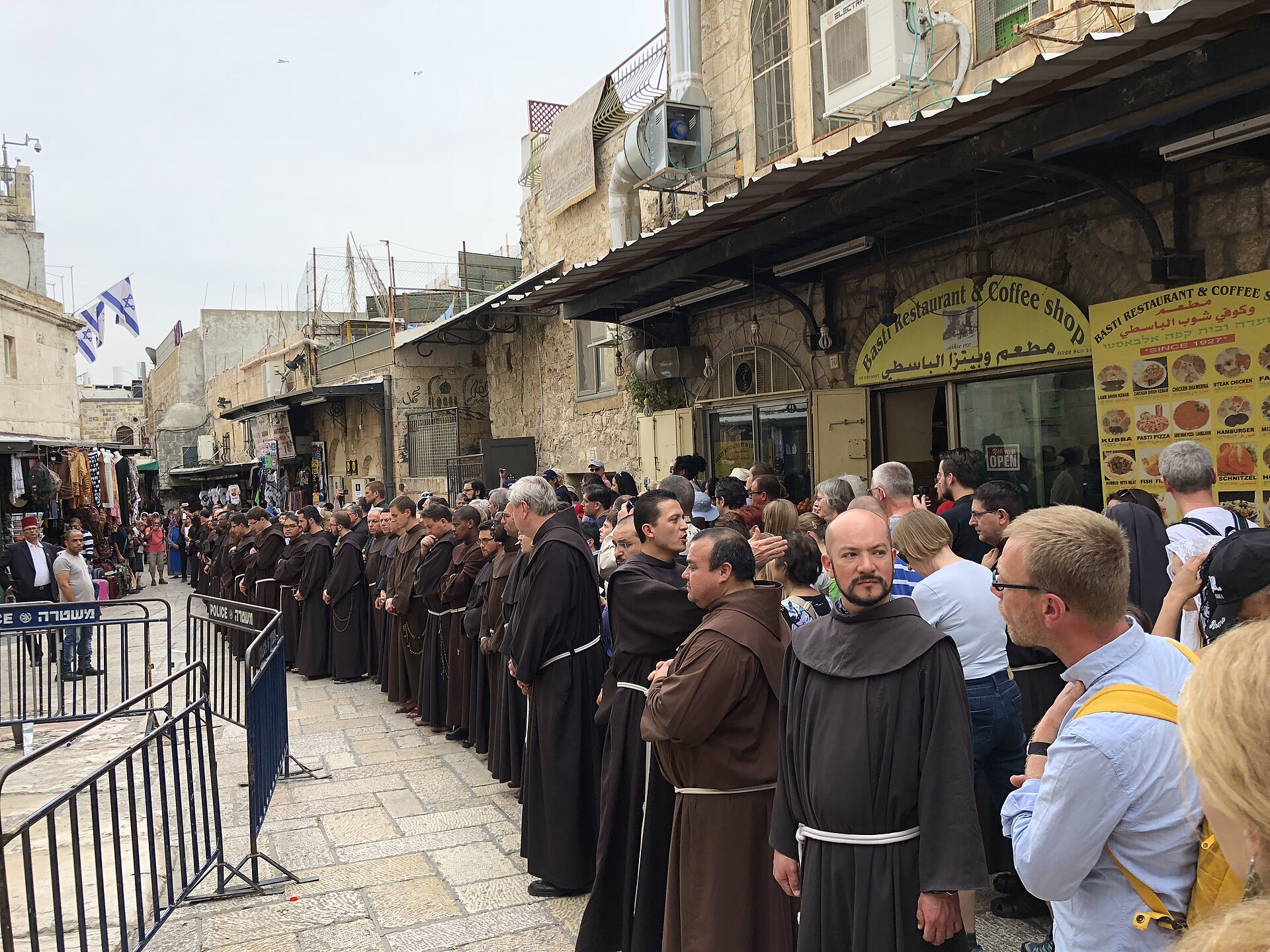 Line of Franciscan friars in brown habits praying along the Via Dolorosa at the Third Station during the weekly Friday procession, with pilgrims and tourists alongside