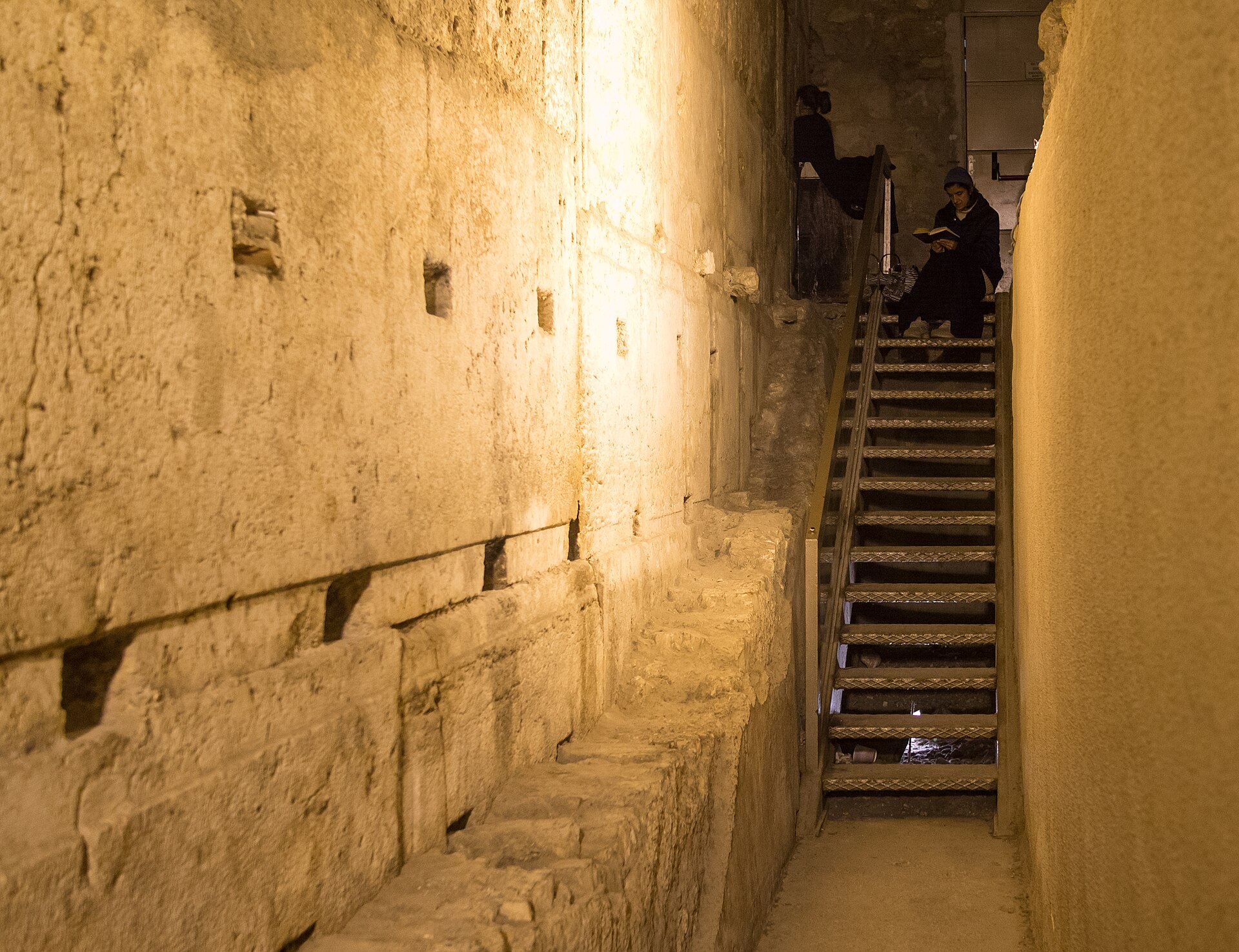 Lit Herodian stone courses of the Western Wall beside a narrow underground passage, where two women read prayer books on a metal staircase