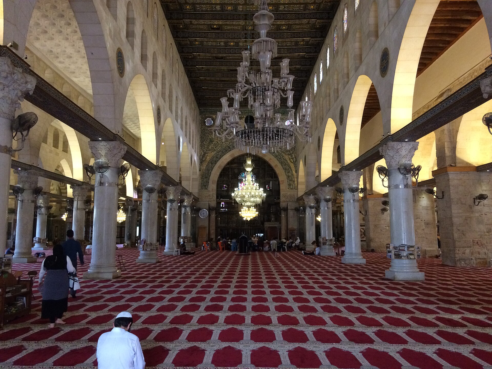 The long central nave of the Al-Aqsa Mosque, with marble columns, pointed arches, crystal chandeliers and red prayer carpets leading to the arch beneath the dome