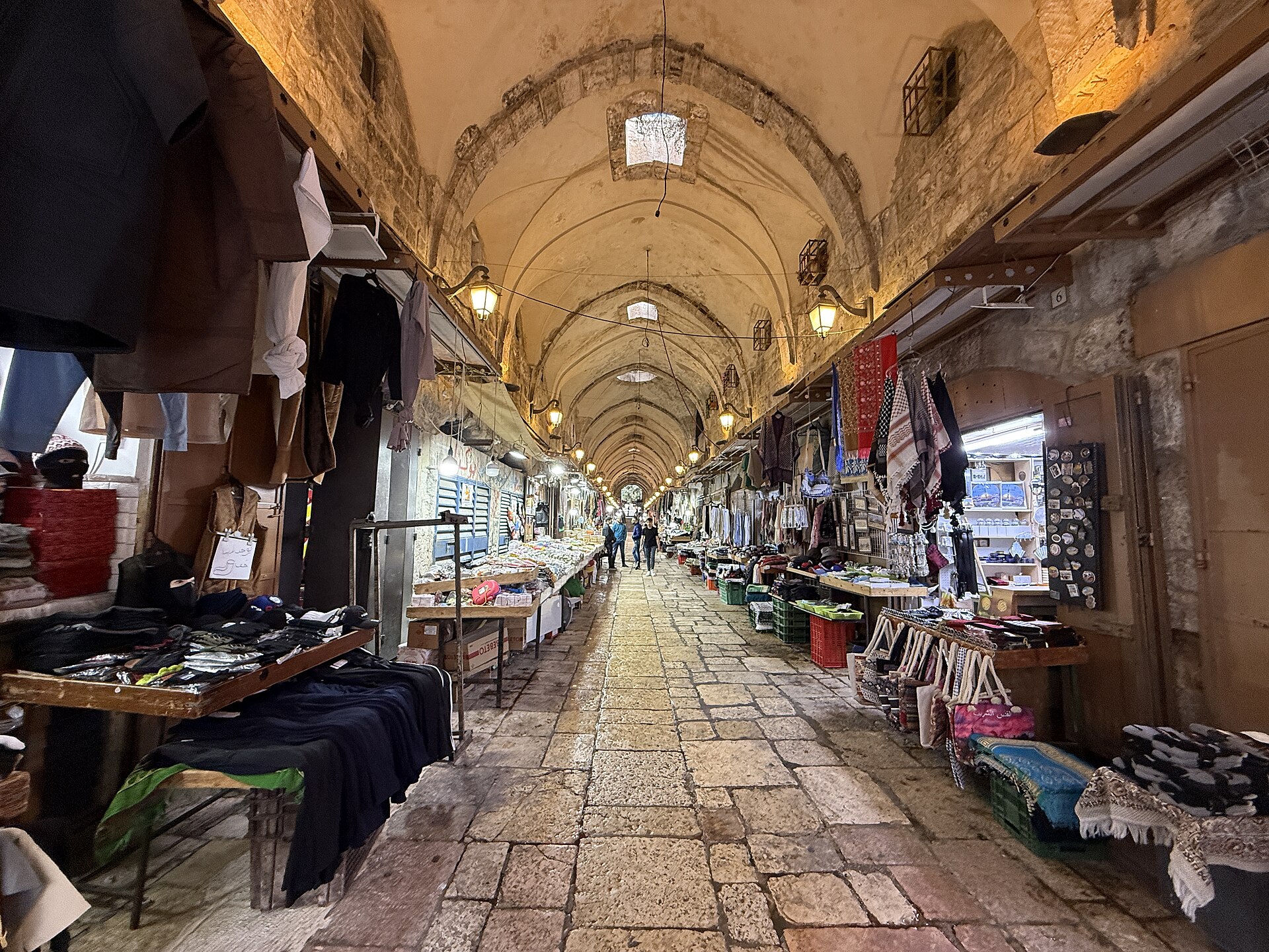 The long vaulted Mamluk arcade of the Cotton Merchants' Market in Jerusalem's Muslim Quarter, lit by lanterns and skylights, with clothing and souvenir stalls