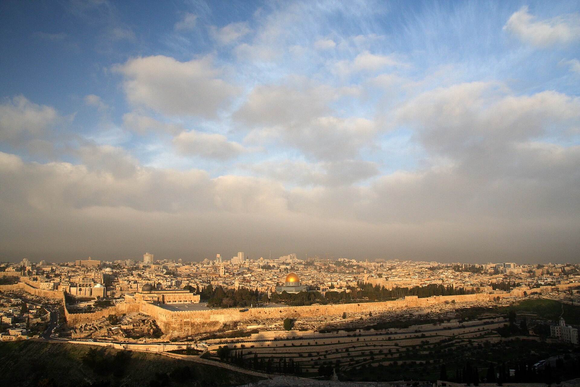 Low early-morning sun lights the Old City walls, Al-Aqsa Mosque and the golden Dome of the Rock beneath a wide cloudy sky, seen from the Mount of Olives