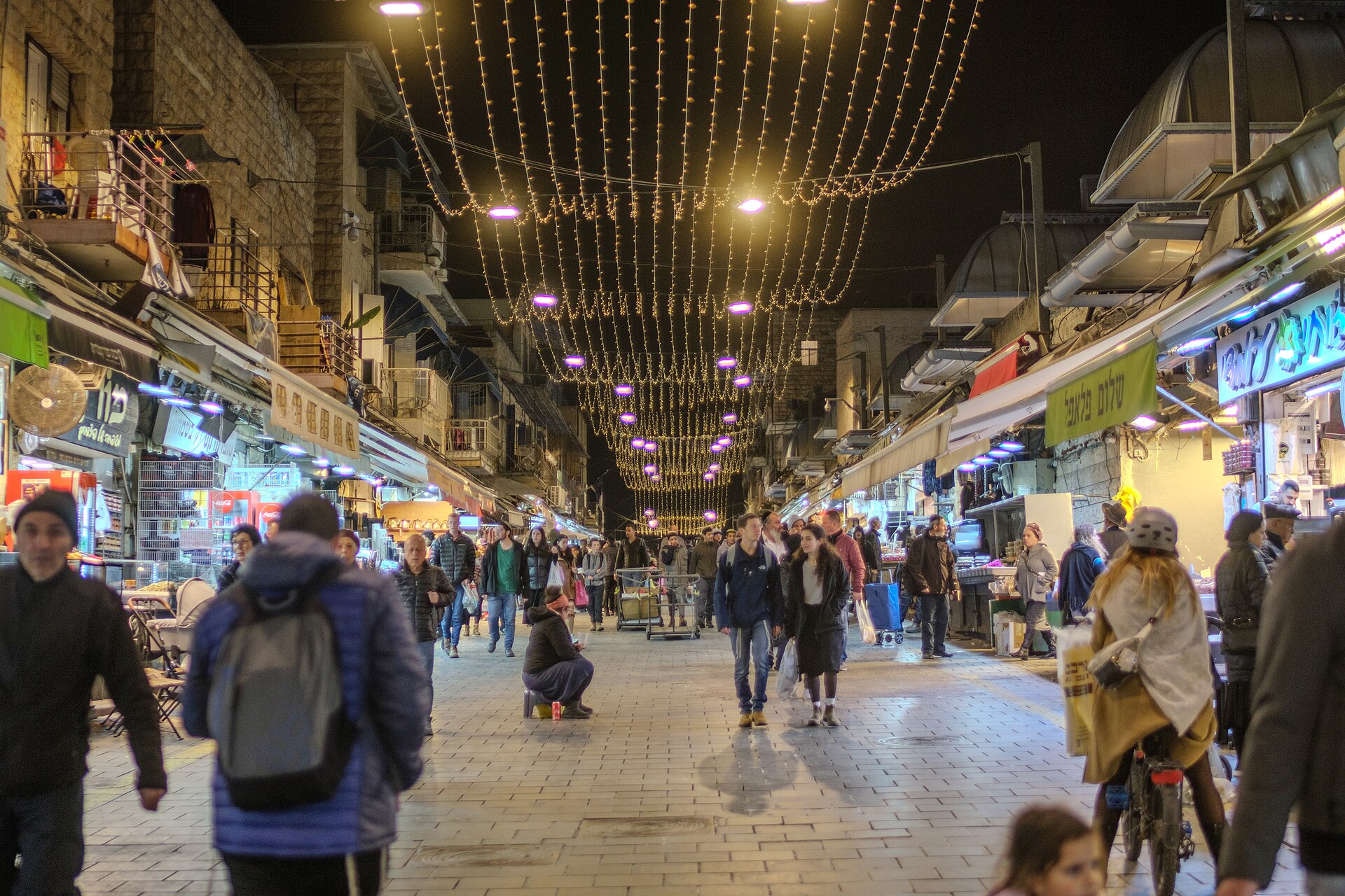 Mahane Yehuda market street at night under canopies of string lights, with shoppers walking between lit stalls and a green Shalom Falafel sign on the right