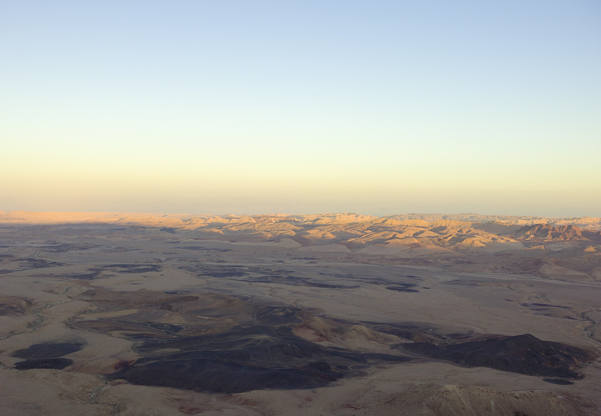 The vast floor of Makhtesh Ramon in the Negev Desert at dawn, with dark volcanic patches and ridges lit pale gold under a clear sky