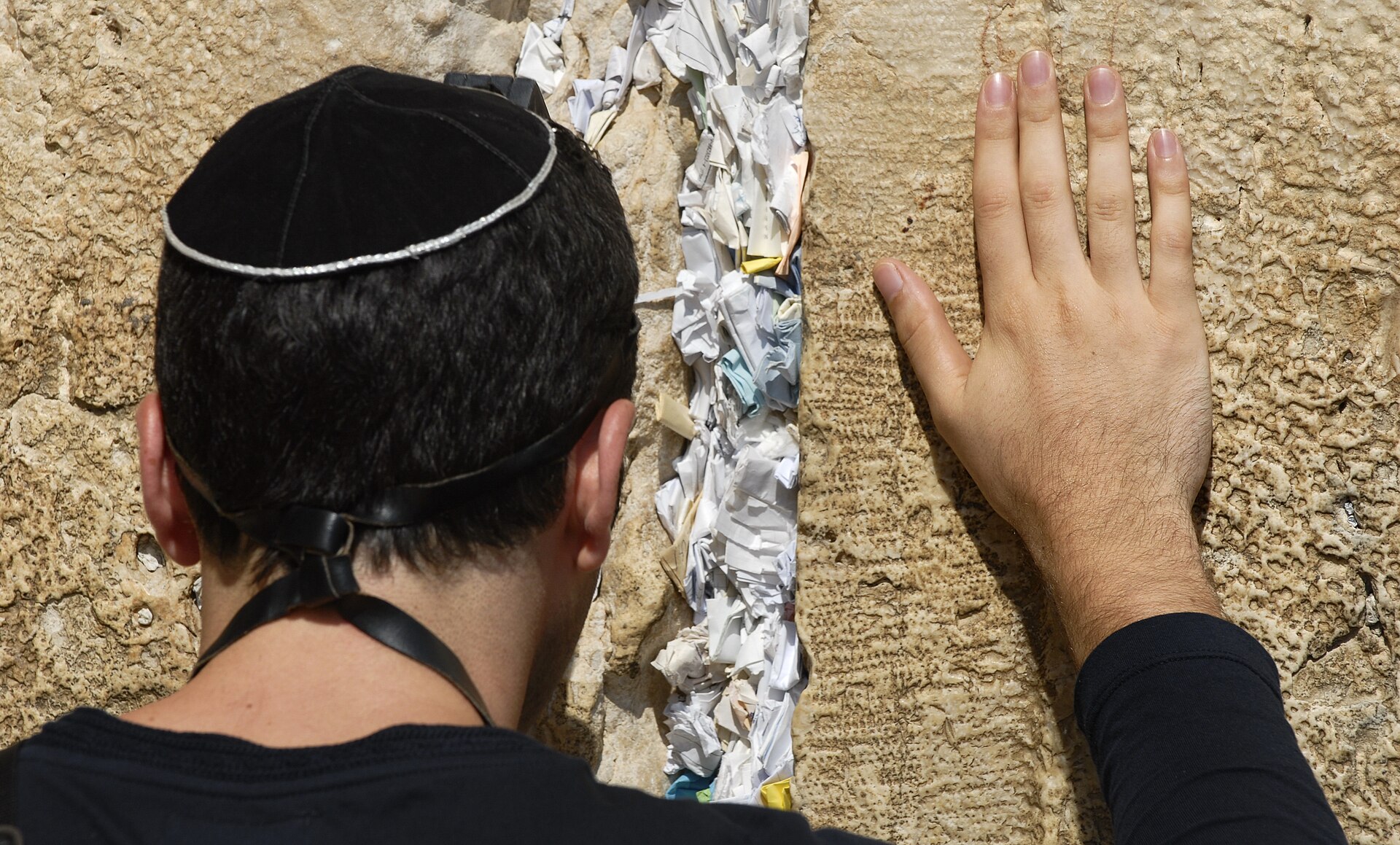 A man in a black kippah and tefillin straps prays with one hand pressed to the Western Wall beside a crack packed with folded paper notes