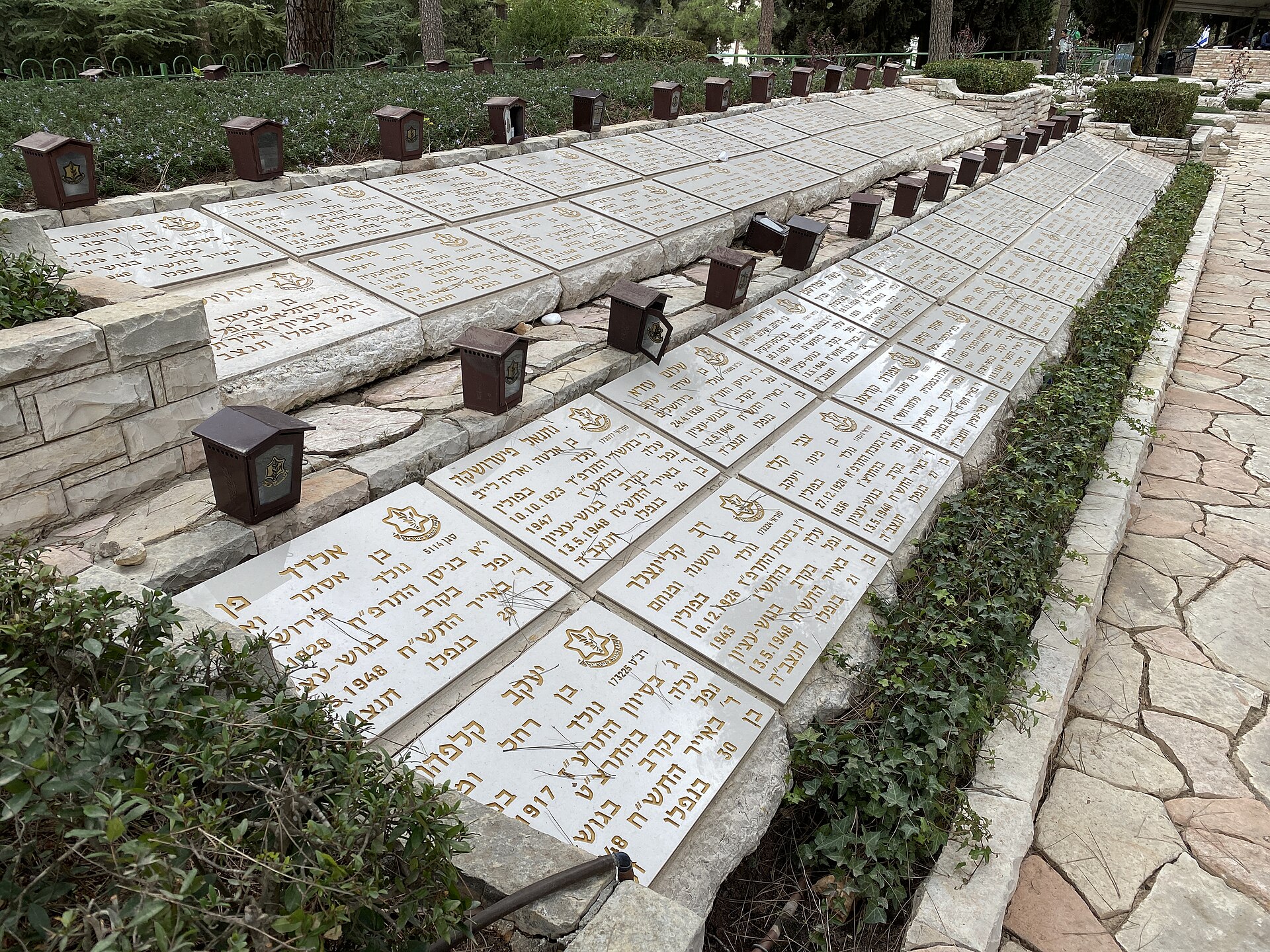 Rows of flat white gravestones with gold Hebrew lettering and army emblems, each with a small memorial lantern, in the military cemetery on Mount Herzl