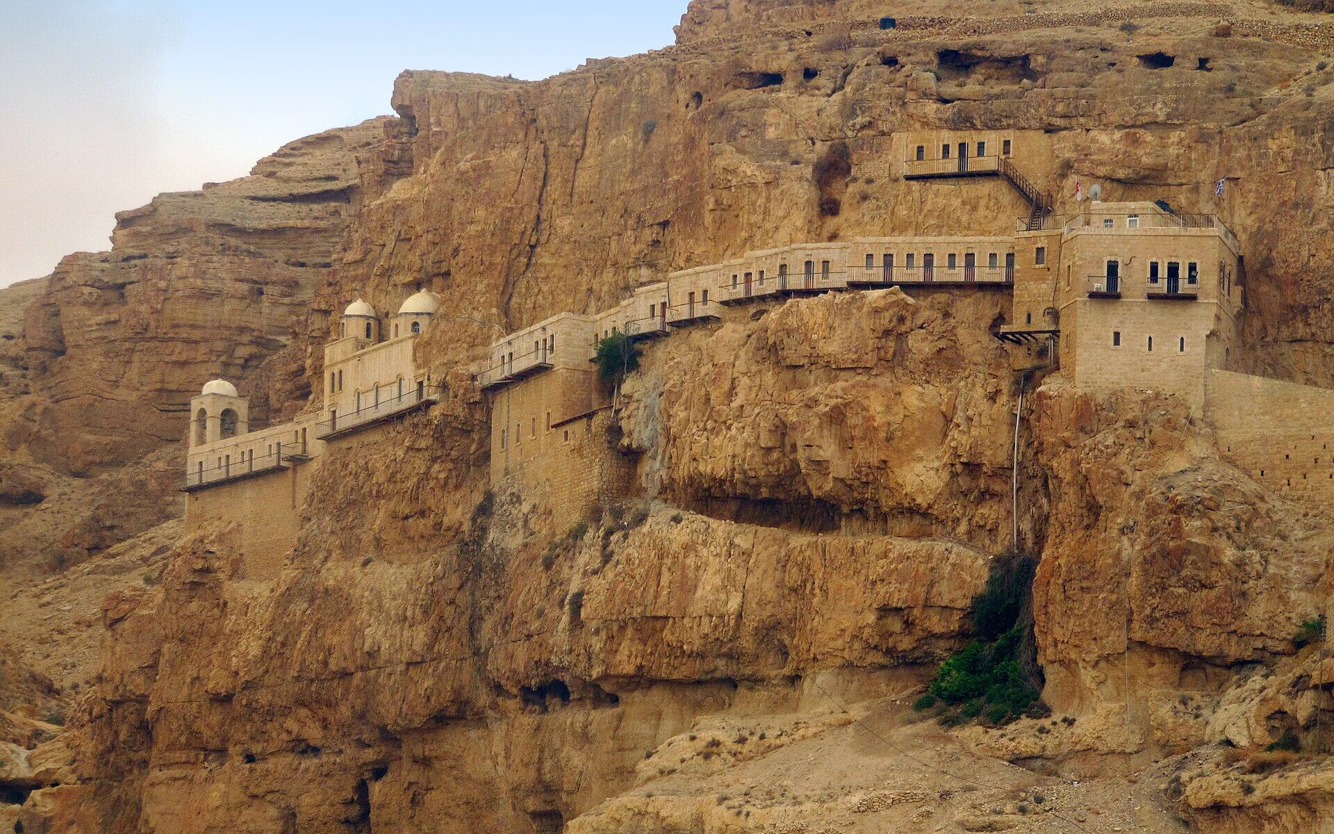 The Monastery of the Temptation, a long line of pale stone buildings with small domes and balconies built along a sheer ochre cliff face above Jericho