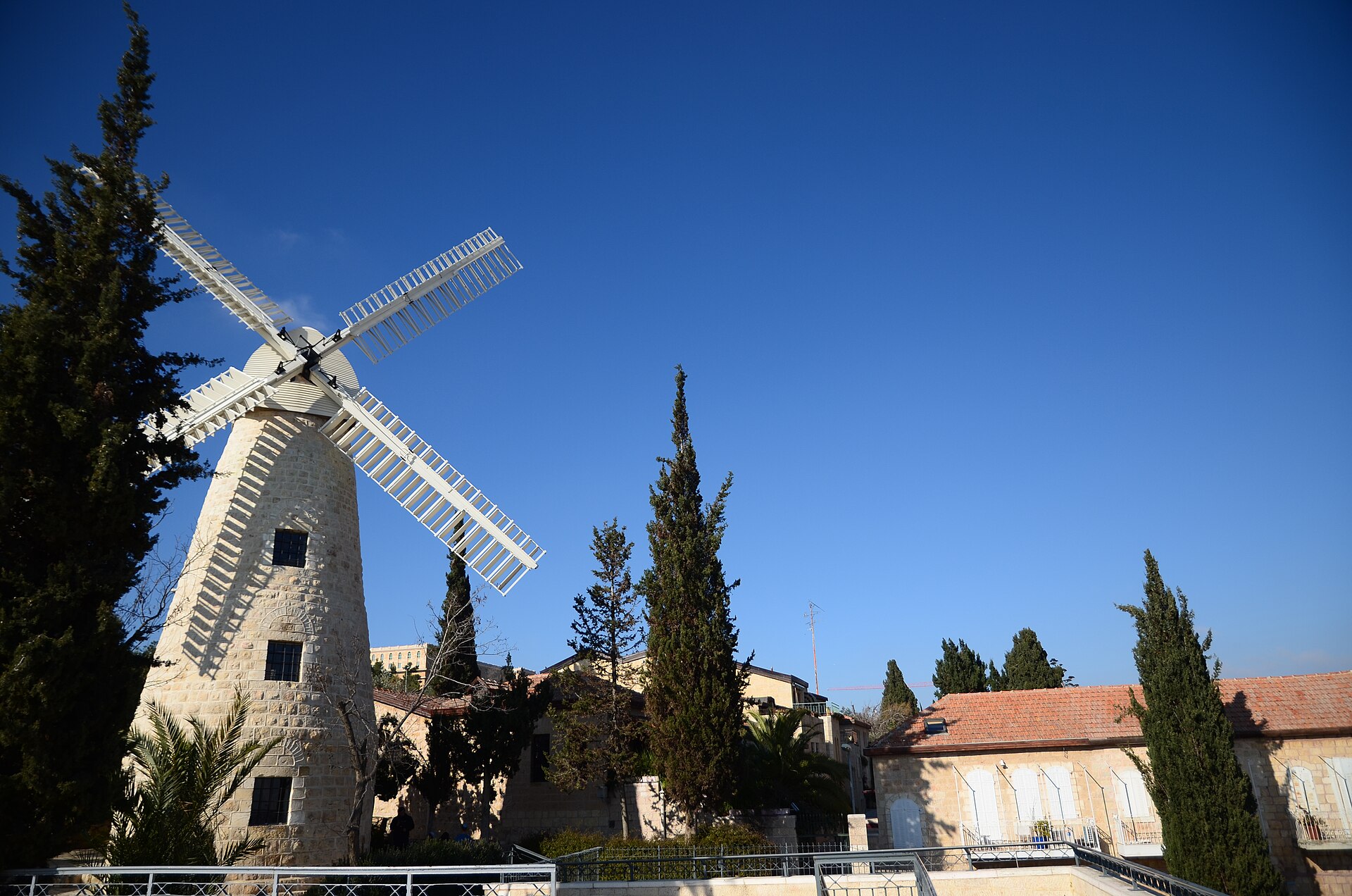 The Montefiore Windmill's stone tower and white sails beside tall cypresses and red-roofed stone houses under a deep blue Jerusalem sky