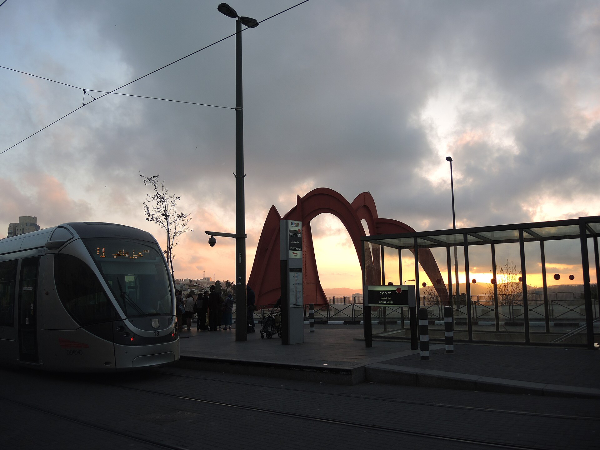 A light rail tram at the Mount Herzl stop at sunset, with a large red Alexander Calder sculpture and a glass shelter beside the platform