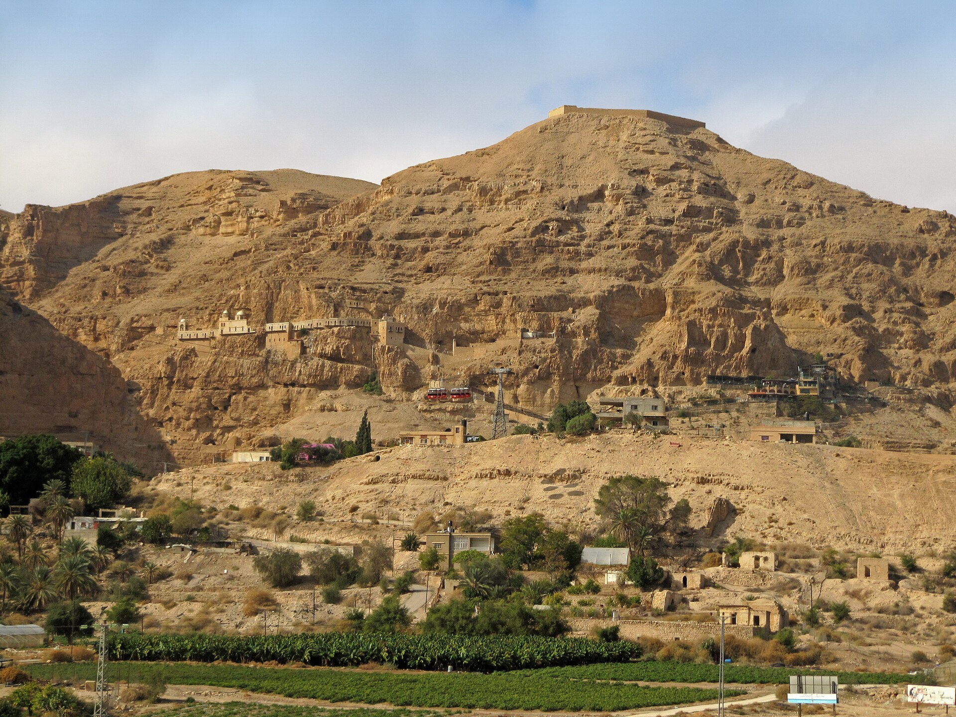 The Mount of Temptation above Jericho, with its monastery on a ledge halfway up the cliff, red cable-car cabins below and a banana plantation in the foreground