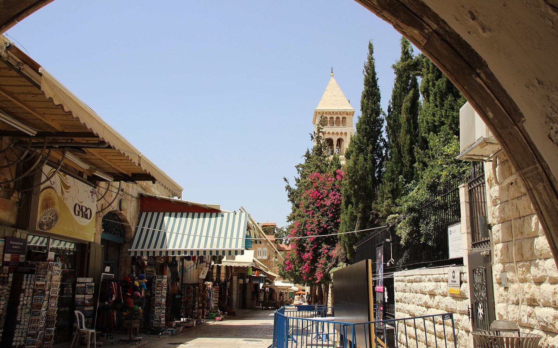 Muristan Road in Jerusalem's Christian Quarter framed by a stone arch, with souvenir shops, cypress trees and the Church of the Redeemer bell tower