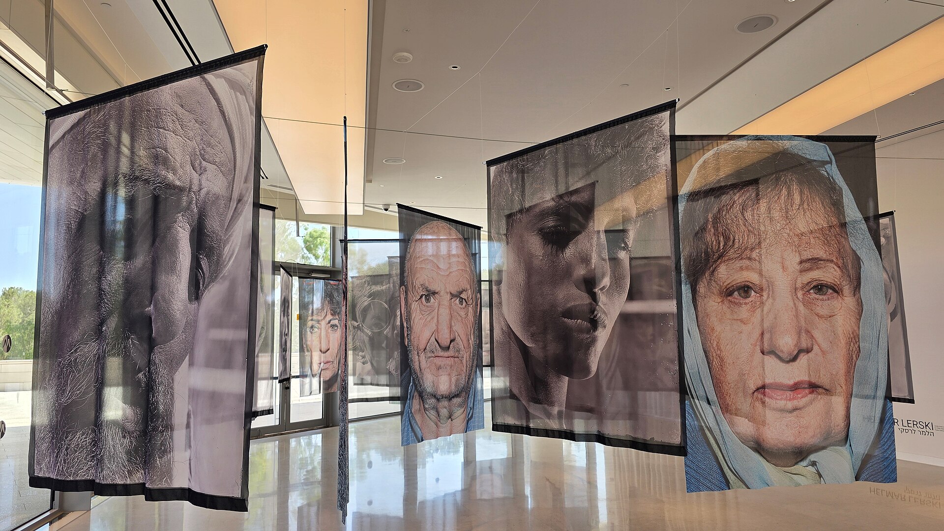 Large translucent fabric banners printed with black-and-white portrait photographs of faces hanging in a bright gallery at the Museum of Tolerance Jerusalem