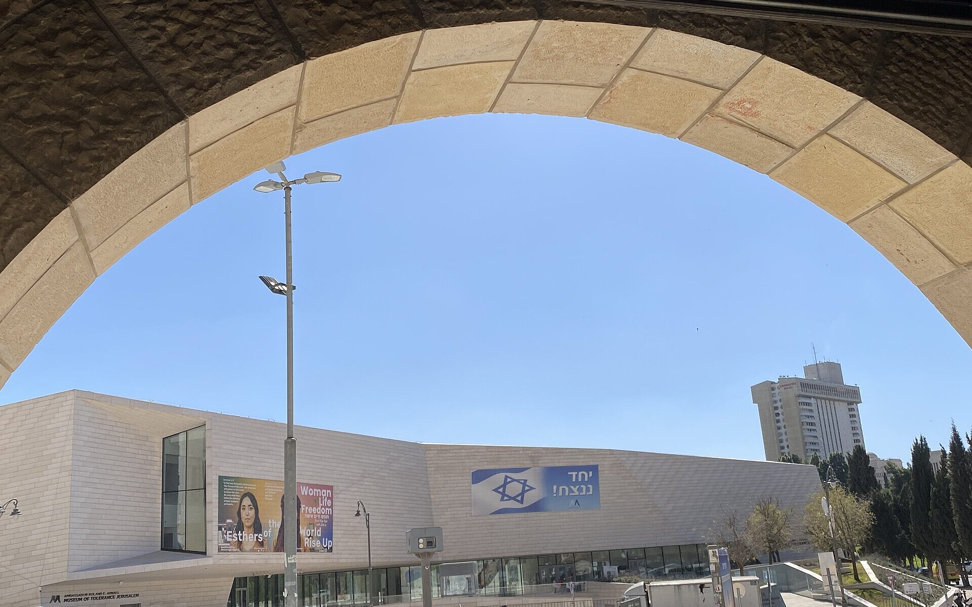 The pale stone facade of the Museum of Tolerance Jerusalem on Hillel Street, seen through a stone archway, with exhibition banners and an Israeli flag banner on its walls