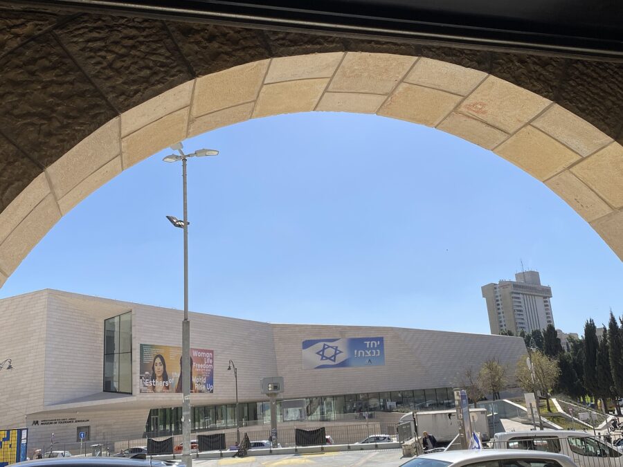The pale stone facade of the Museum of Tolerance Jerusalem on Hillel Street, seen through a stone archway, with exhibition banners and an Israeli flag banner on its walls