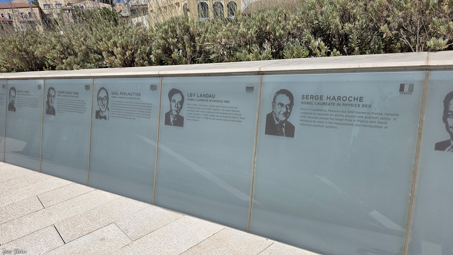 A long glass wall on the terrace of the Museum of Tolerance Jerusalem etched with portraits and short biographies of Nobel laureates in physics, with shrubs and older stone buildings behind