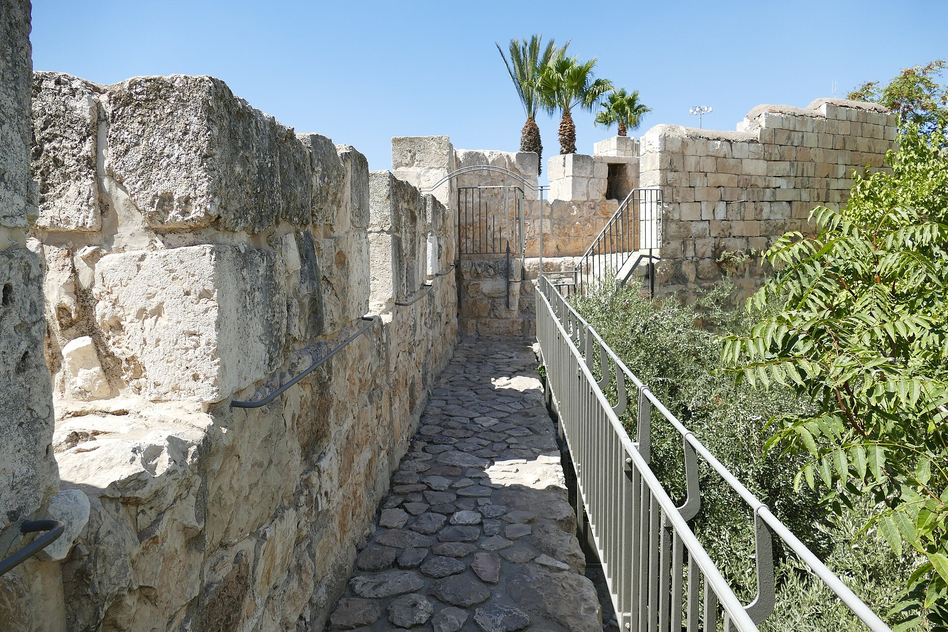 The narrow cobbled Ramparts Walk path on top of Jerusalem's Old City wall, running between the crenellated stone parapet and a metal safety railing towards a tower