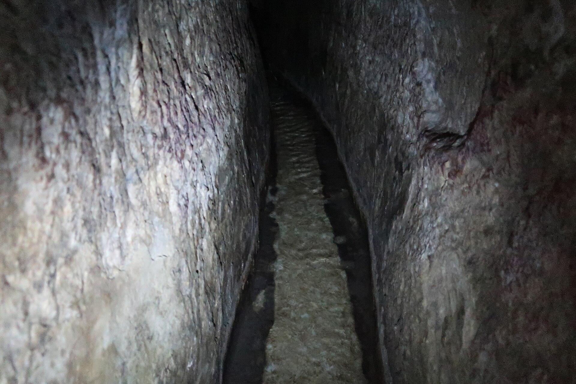 Narrow rock-cut passage of Hezekiah's Tunnel receding into darkness, its torch-lit stone walls rising either side of a shallow stream of water