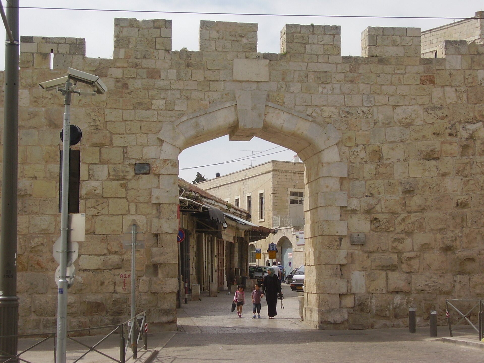 New Gate in Jerusalem's north-western Old City wall, a plain stone arch beneath crenellations, framing a Christian Quarter street with a family walking through