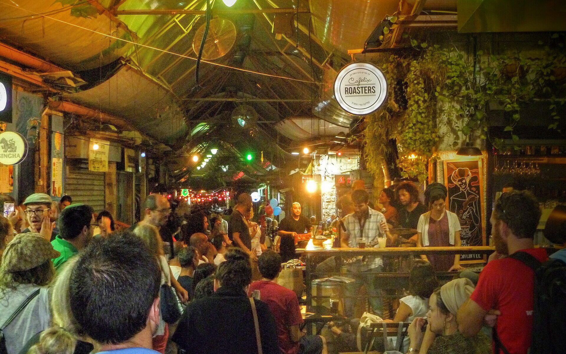 A night crowd packs a covered lane of Mahane Yehuda Market around a bar with a live band, under the round Cafelix Roasters sign, string lights and hanging plants