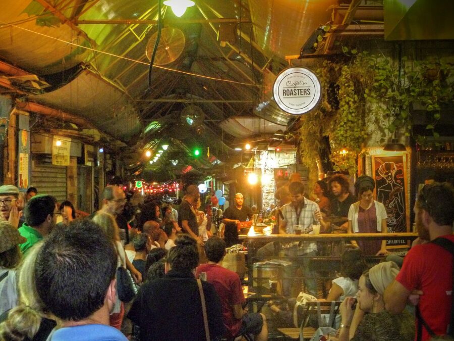 A night crowd packs a covered lane of Mahane Yehuda Market around a bar with a live band, under the round Cafelix Roasters sign, string lights and hanging plants