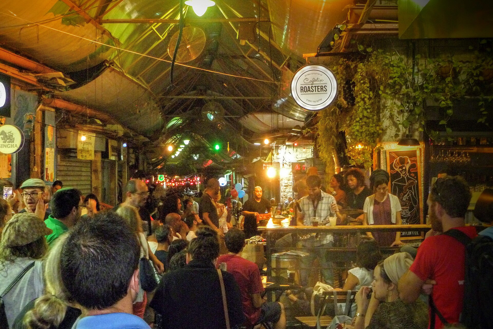 A night crowd packs a covered lane of Mahane Yehuda Market around a bar with a live band, under the round Cafelix Roasters sign, string lights and hanging plants