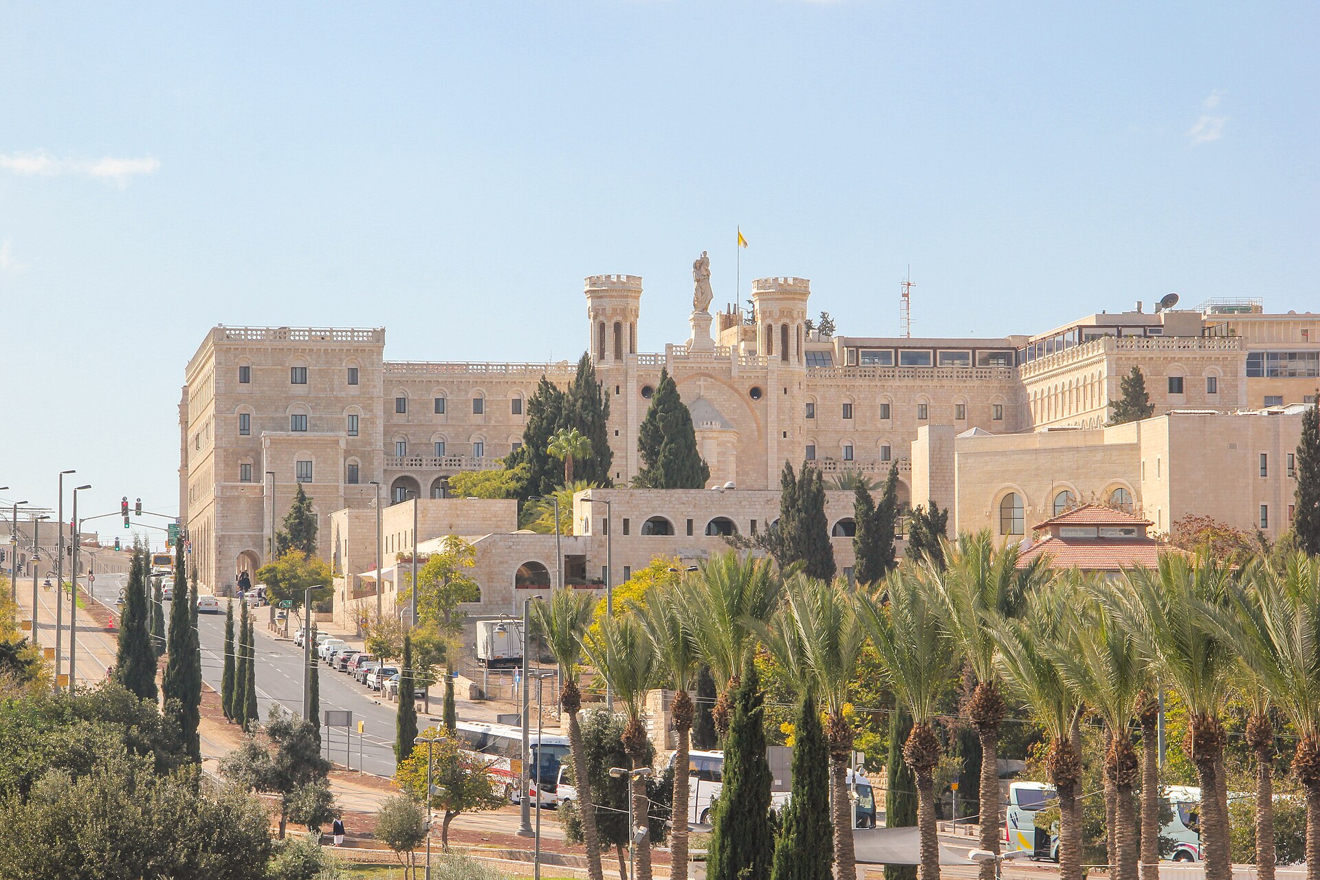 The Notre Dame of Jerusalem Center, a long pale-stone complex with a statue of the Virgin and Child between two crenellated towers, behind rows of palms beside a road