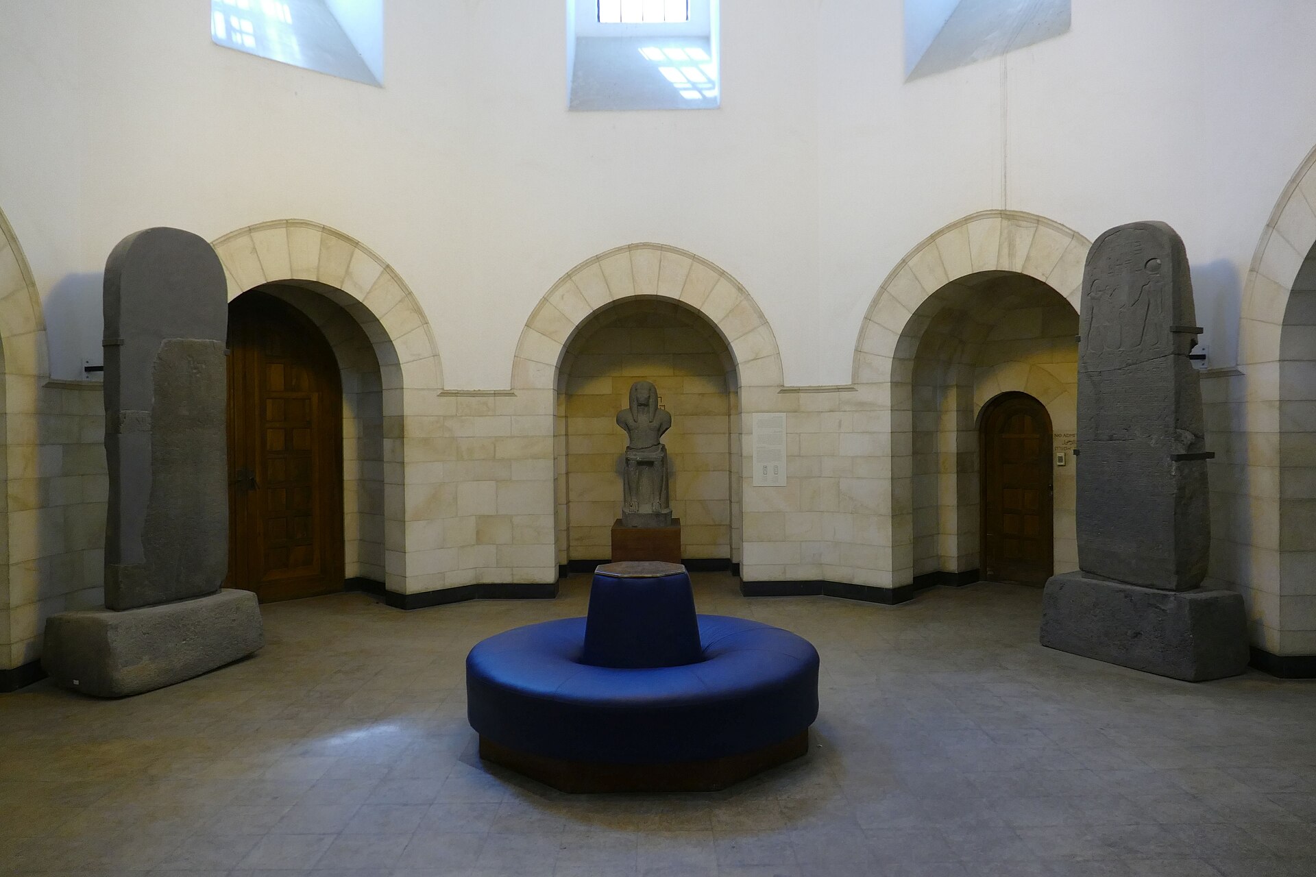 Octagonal gallery of the Rockefeller Museum in Jerusalem, with arched stone niches, two tall basalt stelae, a seated stone statue and a round blue bench