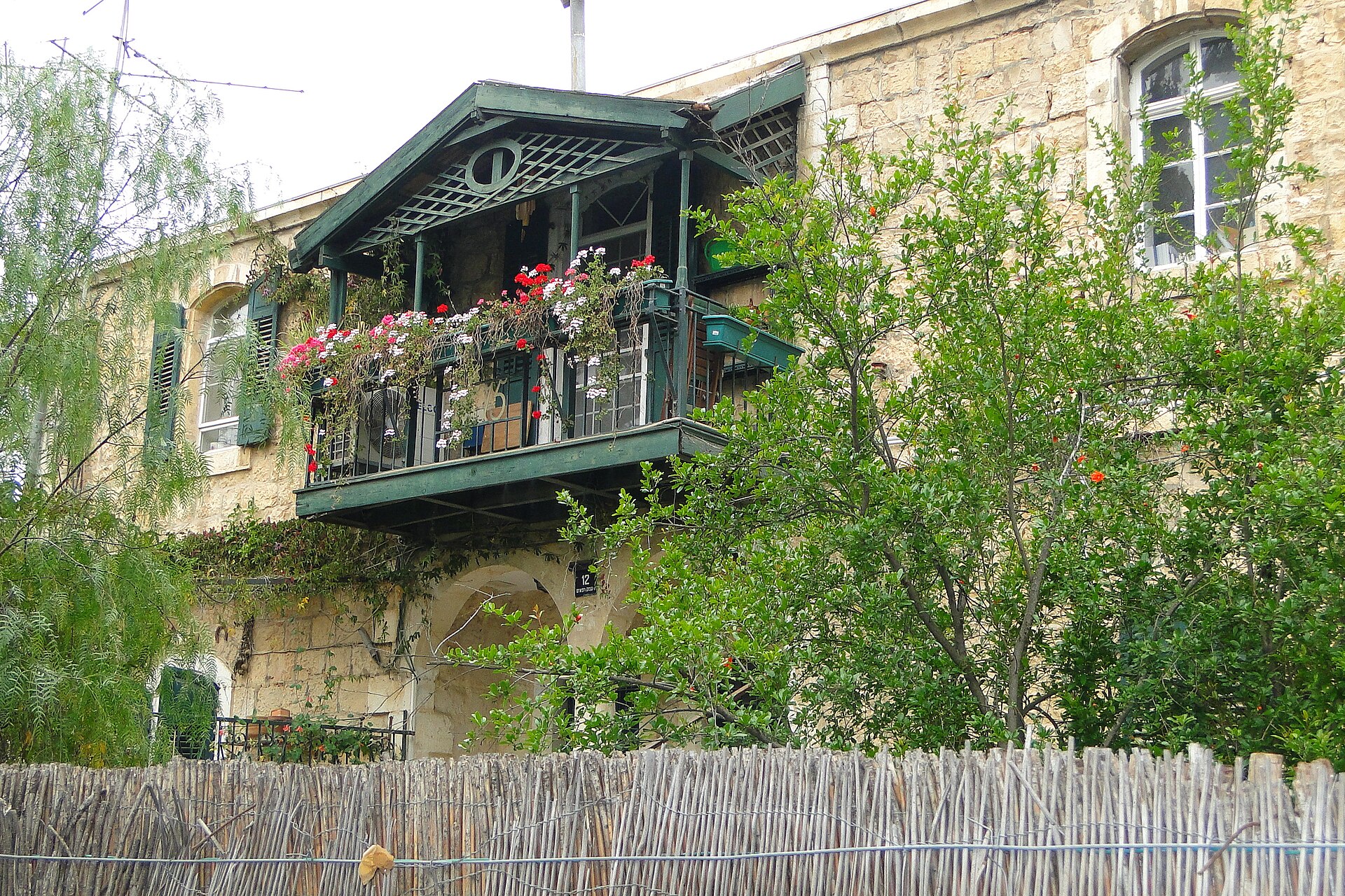 Old stone house at 12 Emek Refaim Street in Jerusalem's German Colony, its green wooden balcony overflowing with geraniums above a reed fence and pomegranate tree