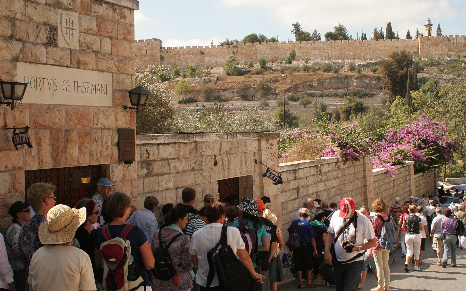 Older tourists in sun hats queue at the Hortus Gethsemani entrance to the Garden of Gethsemane, with bougainvillea on the wall and the Old City walls behind